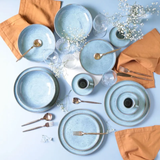 Mesa posta com pratos azuis, talheres dourados, guardanapos laranja e flores brancas