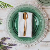 Guardanapo branco dobrado sobre um prato de cristal verde e colher dourada na mesa posta