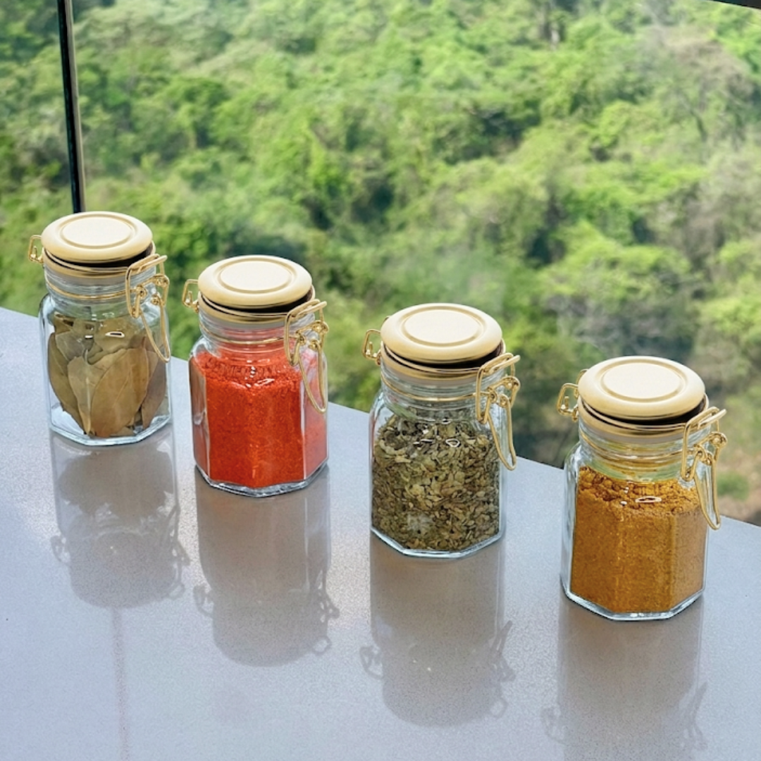 quatro potes de vidro com tampas douradas e temperos coloridos enfileirados