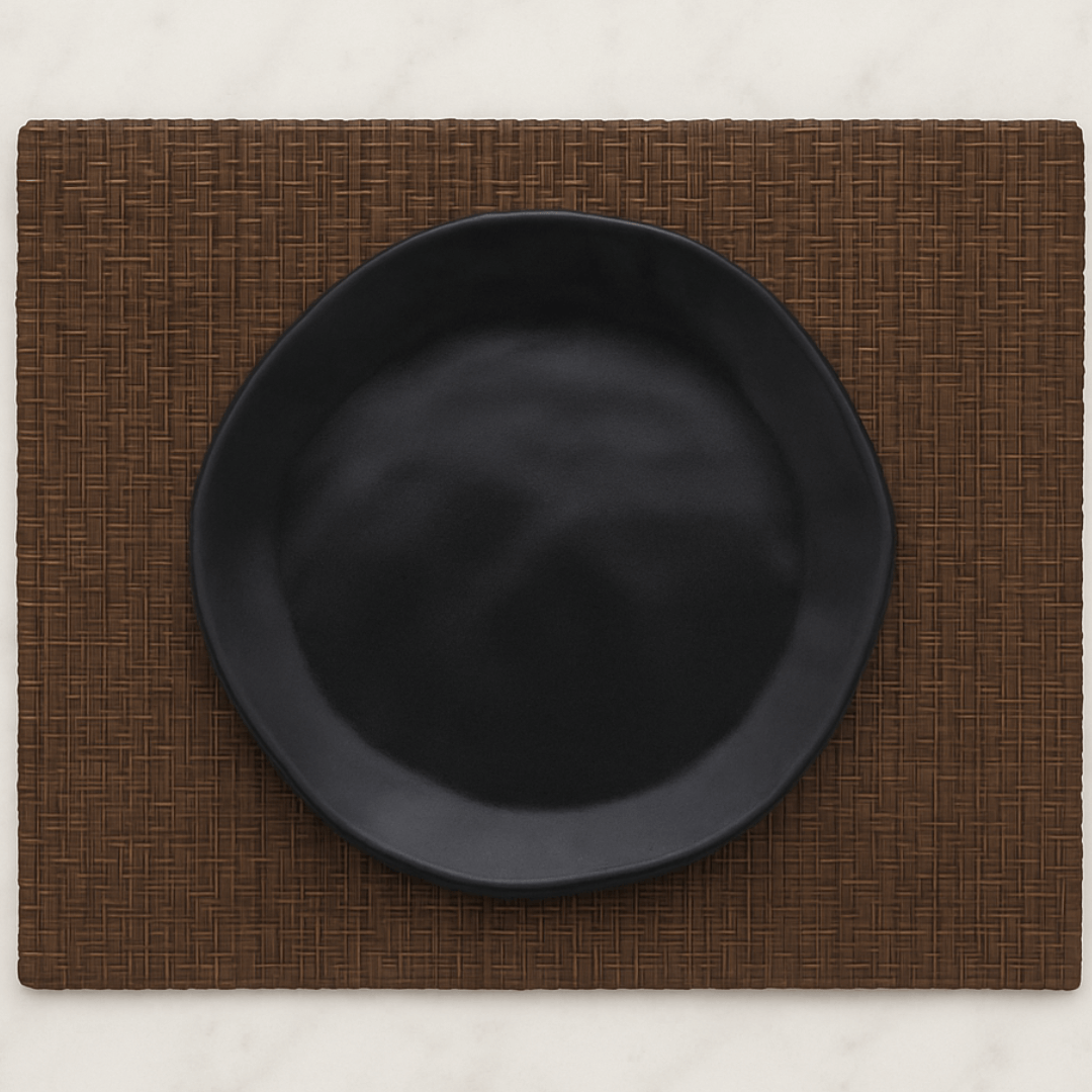 Prato de sobremesa preto com formato orgânico sobre jogo americano marrom, disposto em uma mesa de mármore branco.
