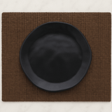 Prato de sobremesa preto com formato orgânico sobre jogo americano marrom, disposto em uma mesa de mármore branco.