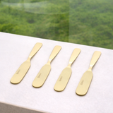 Quatro espátulas aço inox dourado alinhadas em superfície branca