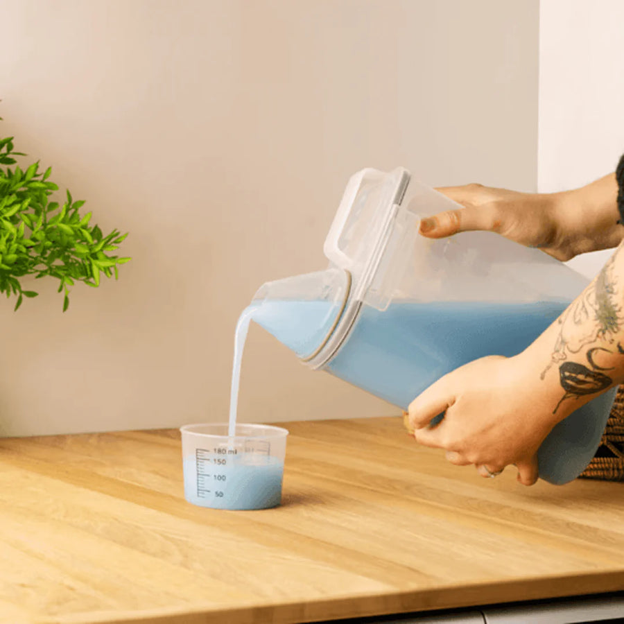 Pessoa despejando sabao liquido azul do pote no copo medidor sobre bancada de madeira