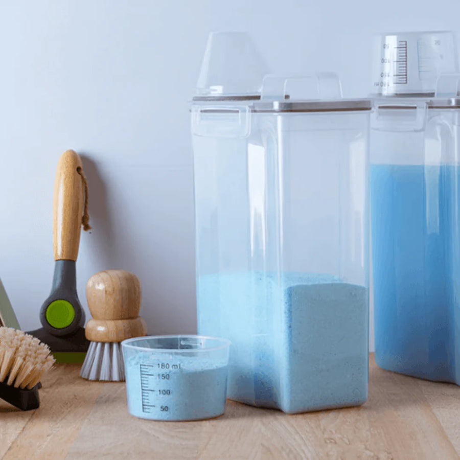 Potes com sabao em po e liquido azul ao lado de escovas sobre bancada de madeira