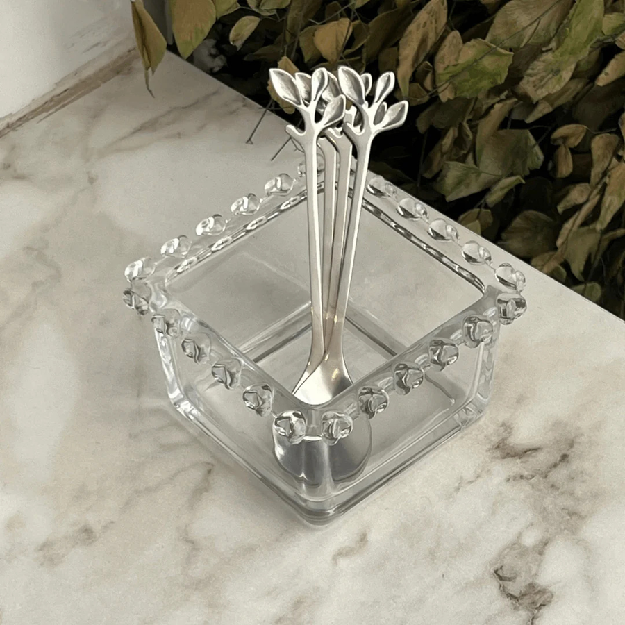 Colheres prateadas com detalhe de folhas apoiadas em bowl quadrado de vidro