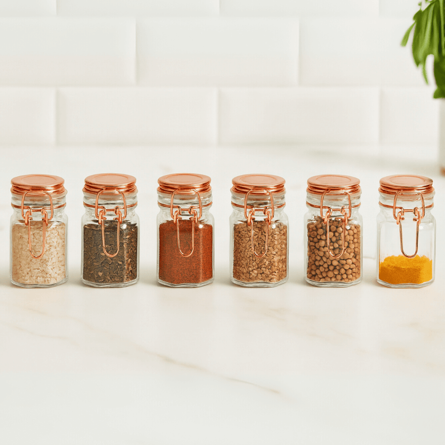 seis potes de vidro herméticos com tampas rose gold enfileirados em bancada branca com temperos variados