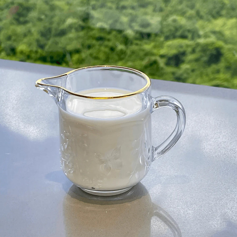 Leiteira de vidro com fio de ouro cheia de leite ao fundo verde natural