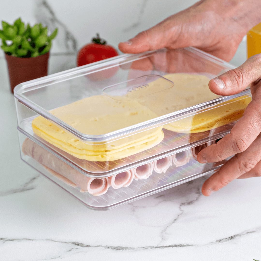 Mãos segurando organizador duplo transparente com queijo fatiado e frios na bancada